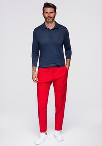 Polo a maniche lunghe blu navy con colletto e logo, abbinato a pantaloni slim-fit rossi e sneakers bianche.