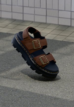 Smeđa kožna sandala s dvije podesive kopče i debelim crnim potplatom, postavljena iznad teksturiranog popločanog poda i pozadine od keramičkih pločica.