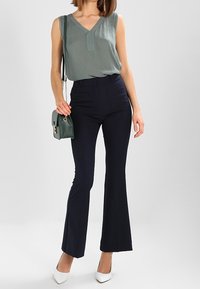 Haut vert sans manches avec un col en V, pantalon taille haute bleu marine, talons blancs et sac à main vert avec une bandoulière en chaîne. Tissus lisses.