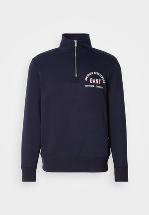 Marineblå hettegenser med høy krage, halv glidelås, ribbede mansjetter og bottom, med hvit tekst som sier "American Sportswear GANT."