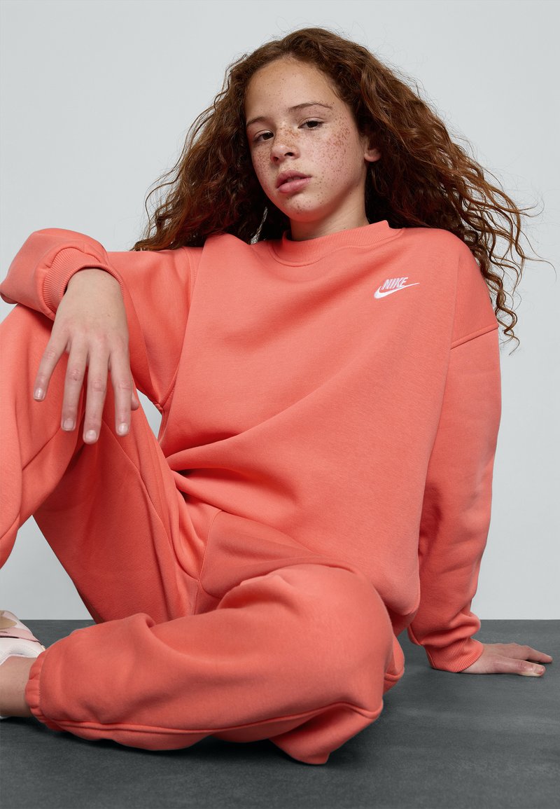 Koralen Nike kapuca z okroglim ovratnikom, sproščenega kroja in belim logotipom. V kombinaciji z ujemajočimi se širokimi trenirkami, obe iz mehke tkanine.