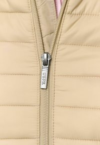 Gai bēšs pūkains mētis ar izšūtu tekstūru. Aprīkots ar sudraba rāvējslēdzēju ar "USPA" zīmolu, iekšpusē redzama kontrastējoša rozā odere.