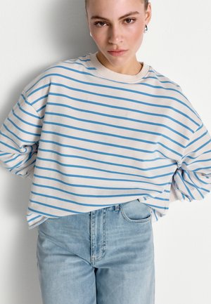 Jauna sieviete ģērbta baltā krekliņā ar gaiši zilām horizontālām svītrām un gaispi ziliem augsta vidukļa džinsiem, pozē pret balto sienu.