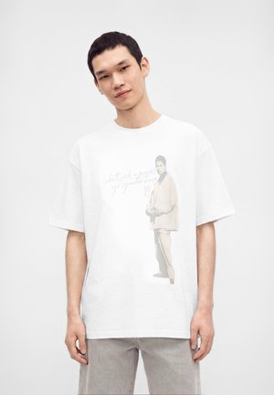 Jonge man met kort donker haar draagt een wit oversized T-shirt met een beige figuurprint en lichtgrijze broek, staand tegen een effen achtergrond.