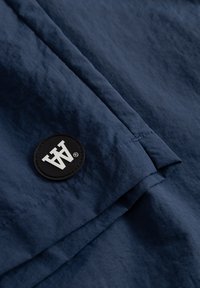 Tissu bleu marine avec une finition texturée, comportant un patch rond noir embossé d'un logo blanc. Lignes épurées et détails de couture subtils.