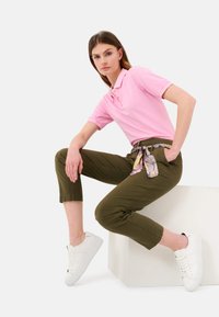Rosa Polohemden mit kurzen Ärmeln, olivgrüne cropped Hose mit einem gemusterten Gürtel und weiße Sneakers an einem Model, das sitzend gegen einen weißen Würfel posiert.