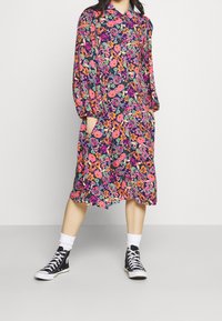 Robe florale avec un fond bleu marine, ornée de grandes fleurs roses et orange. Manches longues et deux poches latérales. Associée à des baskets noires.
