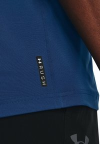 Blue athletic shirt made of smooth fabric, featuring a black label with "RUSH" and Under Armour logo near the bottom.
