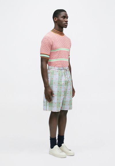 Κοντομάνικο μάλλινο πουλόβερ σε κόκκινο και λευκό με υφή, με πράσινες και μαύρες ρίγες· καρό σορτς με μοτίβα και ανθισμένες λεπτομέρειες· λευκά αθλητικά παπούτσια και ναυτικόσχοινα κάλτσες.