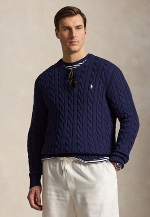 Mężczyzna noszący granatowy sweter w warkocze na paskowanej koszuli, białe spodnie ze ściągaczem, okulary przeciwsłoneczne zawieszone na swetrze i zegarek na lewym nadgarstku.