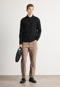 Μαύρο πουκάμισο με κουμπιά, καφέ παντελόνι με λεπτομέρειες από μπαλώματα και μαύρα παπούτσια; κρατά μία μαύρη τσάντα. Λείο ύφασμα, προσαρμοσμένη εφαρμογή.