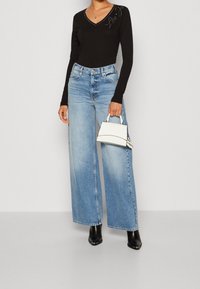 Donna con maglia nera a maniche lunghe, jeans a palazzo blu chiaro, stivali neri, che tiene una piccola borsa bianca contro uno sfondo semplice.