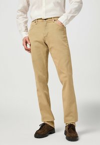 Licht tan katoenen chinos met een rechte pijp, voorzien van vijf zakken en zichtbare stiksels. Gecombineerd met donkerbruine veterschoenen.