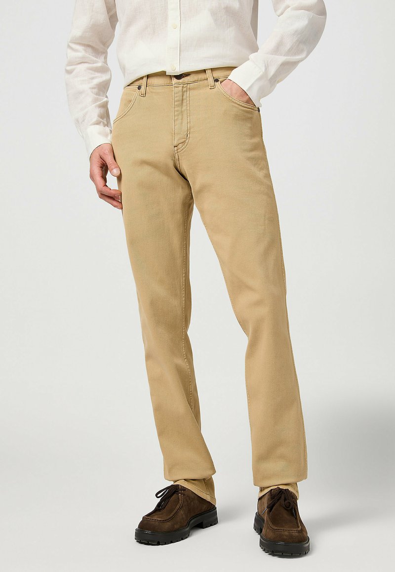 Licht tan katoenen chinos met een rechte pijp, voorzien van vijf zakken en zichtbare stiksels. Gecombineerd met donkerbruine veterschoenen.