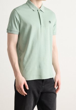 Man wearing short-sleeve sage green polo shirt with small chest logo paired with black pants, standing against plain background.