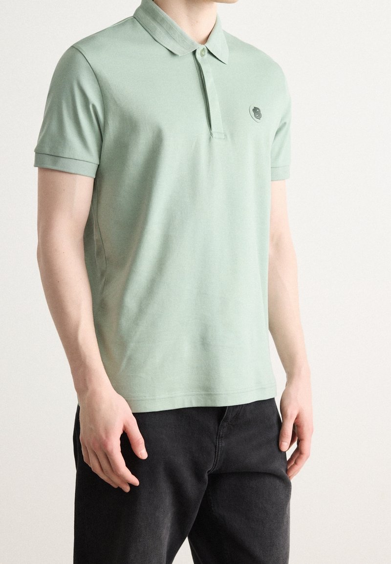Homme portant un polo vert sauge à manches courtes avec un petit logo sur la poitrine, associé à un pantalon noir, debout devant un fond uni.