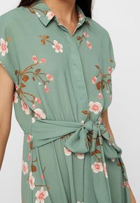 Robe vert clair à manches courtes, motif floral en rose et blanc, col chemise, et détail de taille nouée. Texture de tissu douce et fluide.