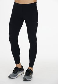 Zwarte leggings van rekbare stof, met een platte tailleband, zijzakken en subtiele stiksels. Gecombineerd met grijze sportschoenen.