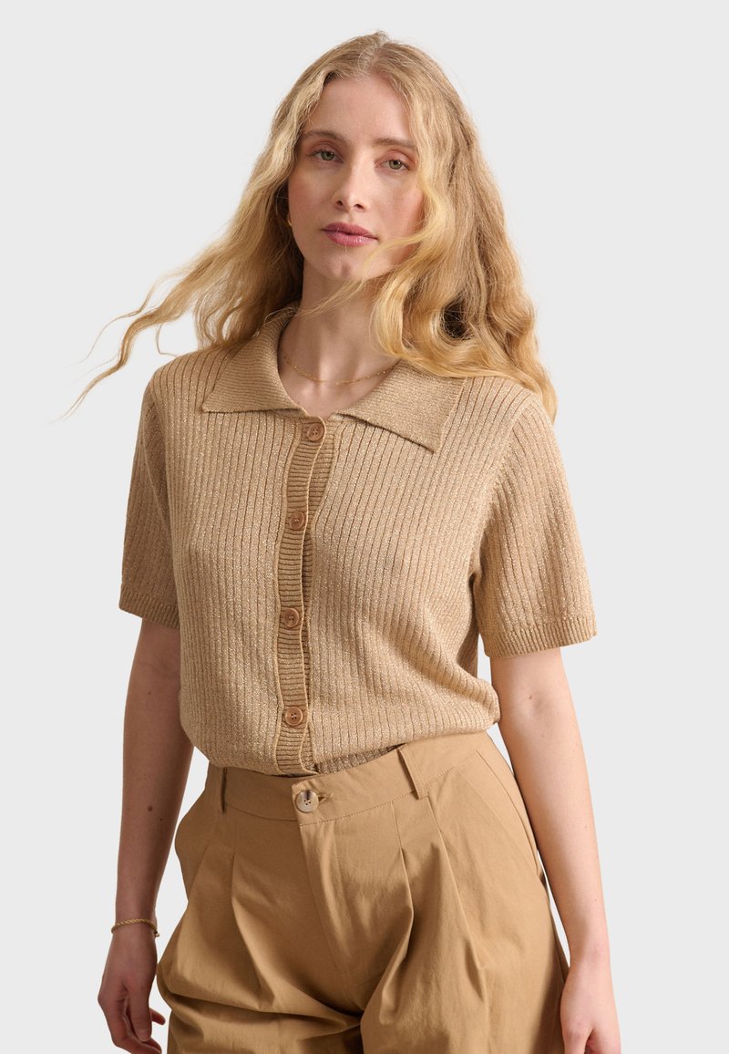 Cardigan beige à manches courtes avec un col évasé et cinq boutons marron sur le devant. La texture est douce et légèrement scintillante.