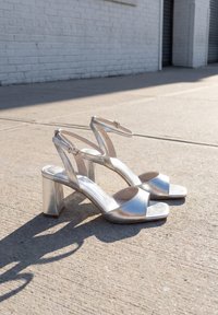 Zilverkleurige sandalen met hoge hakken en enkelbandjes, geplaatst op een zonovergoten betonnen stoep naast een witte bakstenen muur.