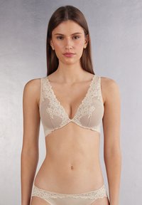Béžová krajková podprsenka s hlubokým V-výstřihem, zdobená květinovou krajkou na ramínkách a košíčcích, s texturovaným materiálem v jemném provedení.