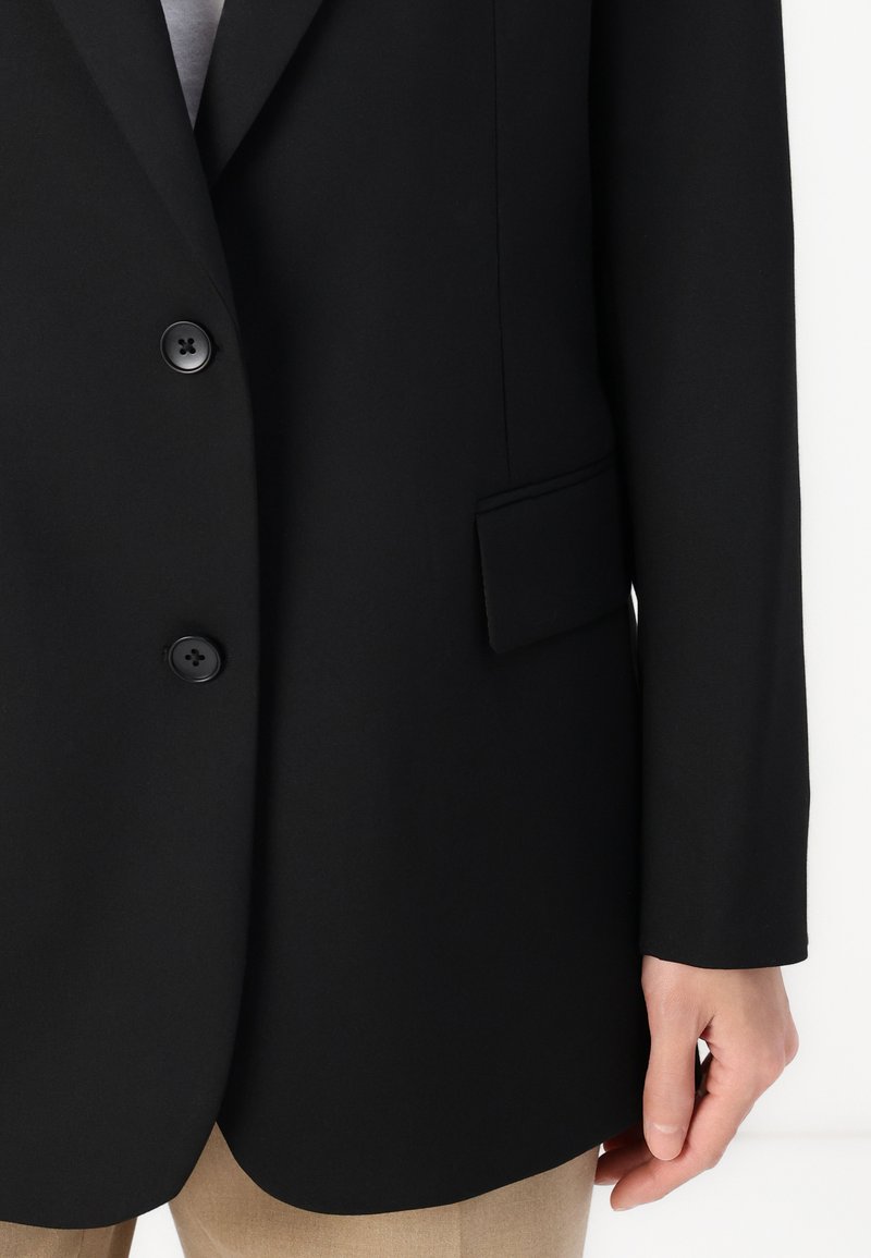 Schwarzer Blazer mit zwei Knöpfen, einer einzelnen Brusttasche und glattem Stoff. Lange Ärmel und taillierte Passform. Über beigen Hosen getragen.