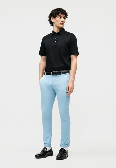 Νέος άνδρας με σγουρά μαλλιά, που φοράει μαύρο κοντομάνικο πόλο, ανοιχτόχρωμο γαλάζιο στενό παντελόνι, μαύρη ζώνη και μαύρα παπούτσια, όρθιος με τα χέρια στις τσέπες.