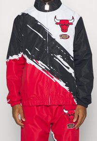 Rød- og sort vindjakke med hvide skrå striber, featuring et Bulls-logo og NBA Finals-patch. Glat, syntetisk materiale; elastiske manchetter.