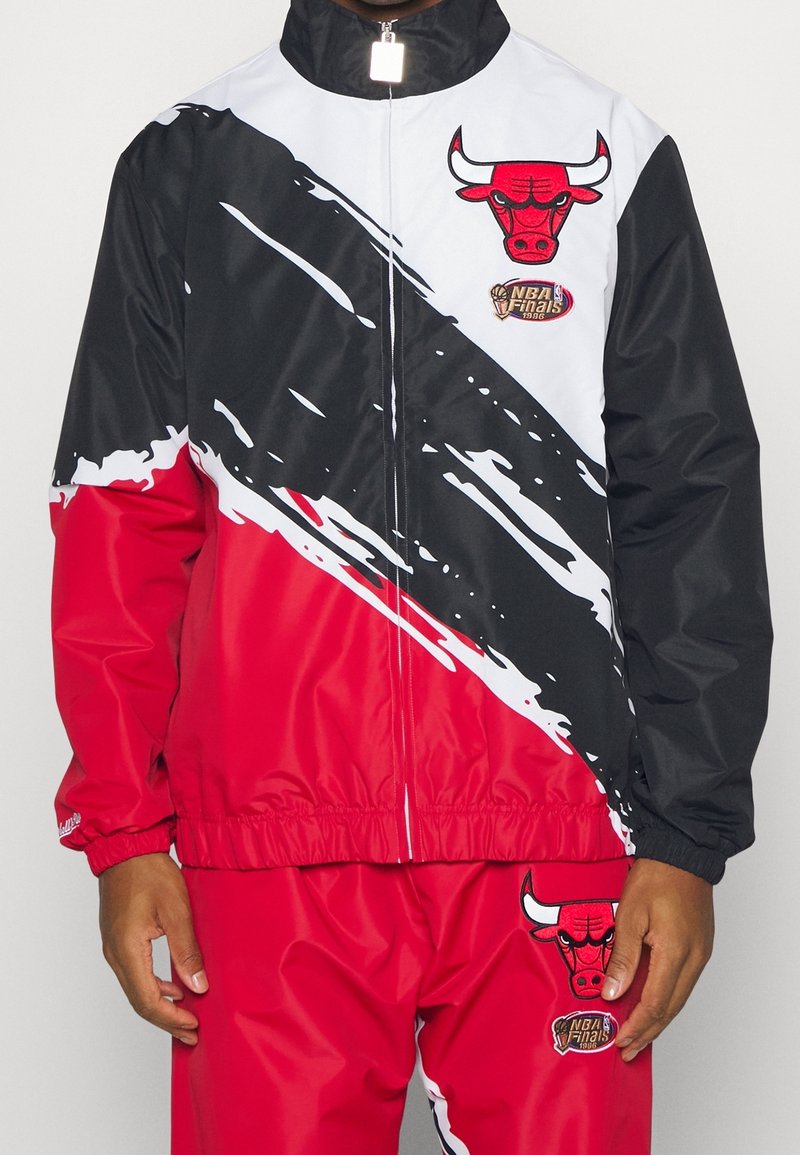 Rød- og sort vindjakke med hvide skrå striber, featuring et Bulls-logo og NBA Finals-patch. Glat, syntetisk materiale; elastiske manchetter.