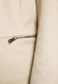 Beige tyg med fin ribbning, med en synlig metalldragkedja på sidan som ger en funktionell detalj till plagget.