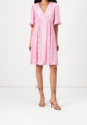 Mujer con un vestido rosa hasta la rodilla con mangas cortas abullonadas y escote en v, combinado con sandalias de tacón alto de tiras color beige.