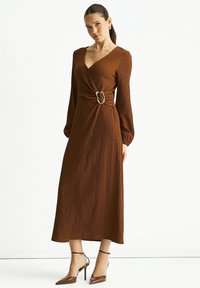 Brown wrap dress with long sleeves, textured fabric, cinched waist featuring a metallic buckle, and ankle-high pointed heels.