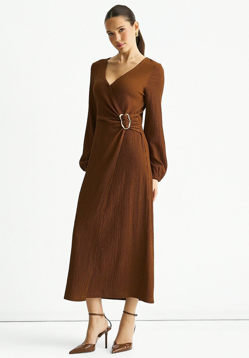 Brown wrap dress with long sleeves, textured fabric, cinched waist featuring a metallic buckle, and ankle-high pointed heels.
