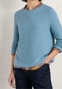 Lichtblauwe textuursweatshirt met lange mouwen en een ronde hals, gedragen over een geruit overhemd met warme kleuren aan de manchetten.