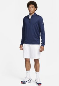 Camisa de manga longa azul-marinho com fecho éclair e um padrão texturizado, com uma gola polo branca por baixo, combinada com calções brancos e sapatilhas multicoloridas.