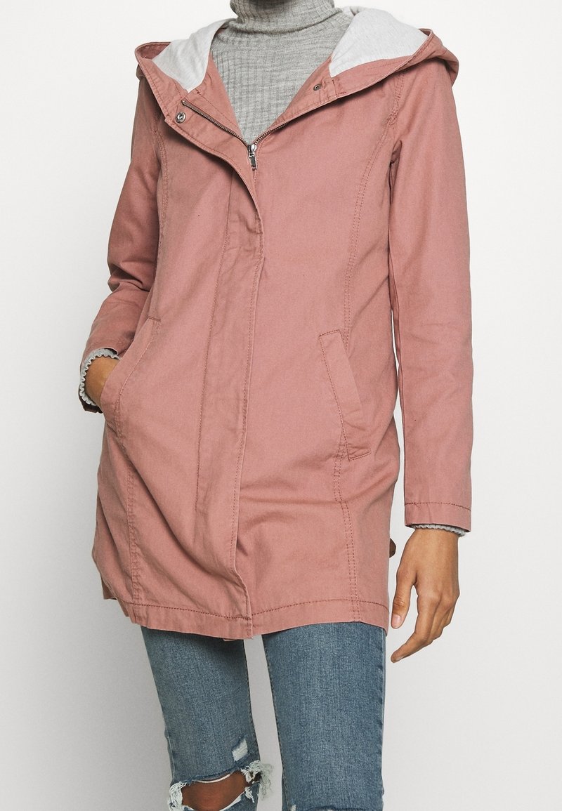 Růžová kapucňová bunda se zipem vpředu, postranními kapsami a šedým podšívkou. Má mírně zakřivený spodní lem a jemné švy.