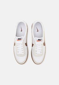 Ténis Nike brancos com atacadores planos, caixa de biqueira semelhante a couro, detalhes em bege, swoosh castanho e sola de borracha texturizada. Logótipo da marca visível.