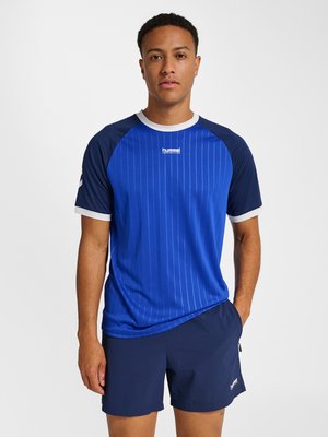 Man staat in een blauw sportshirt met korte mouwen met witte bies en bijpassende marineblauwe short tegen een effen achtergrond.