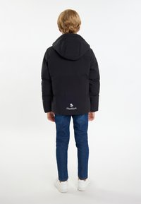 Svart vadderad jacka med huva, med en logotyp på baksidan och elastiska ärmslut, tillsammans med blå jeans och vita sneakers.