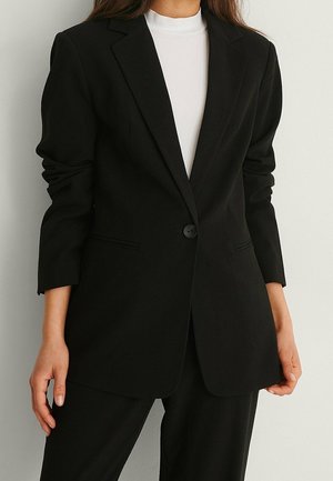 Kobieta w czarnej, dopasowanej marynarce zapinanej na jeden guzik, na białym topie z wysokim kołnierzem, stojąca na jasnym, jednolitym tle.