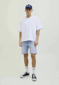 Hvit oversized kortermet skjorte, lyseblå denimshorts, hvite sokker, svarte joggesko, og en blå caps med hvit tekst.