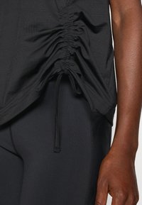 Haut de sport noir avec des détails froncés sur les côtés et un cordon de serrage réglable. Associé à des leggings noirs élégants, montrant une texture et un ajustement lisses.