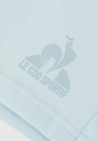 Camisa de algodón azul clara con un estampado sutil que presenta el logo "LE COQ SPORTIF". Textura suave y diseño simple con costuras cosidas.