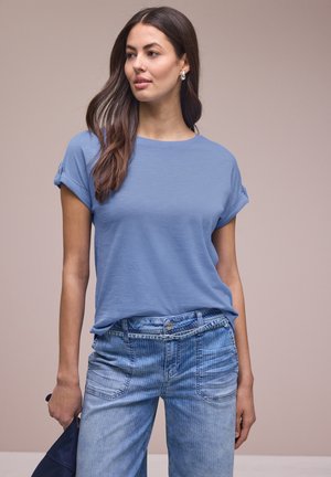 Frau mit langen braunen Haaren, die ein hellblaues T-Shirt mit hochgekrempelten Ärmeln und hellblaue Jeans trägt, hält einen dunkelblauen Gegenstand in ihrer linken Hand.