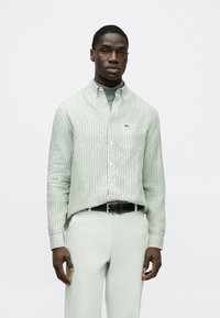 Πουκάμισο με κουμπιά σε πράσινες και λευκές ρίγες, με διπλωμένο γιακά και μικρό λογότυπο. Φορεμένο με παντελόνι ανοιχτού χρώματος και μαύρη ζώνη.