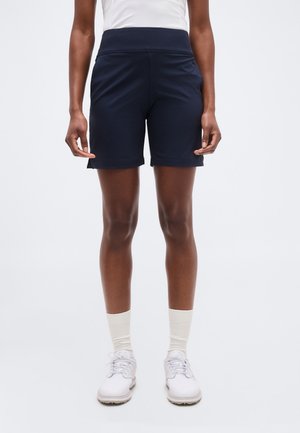 Άτομο που φοράει αθλητικό σορτς σε σκούρο μπλε χρώμα, λευκές κάλτσες πλήρους μήκους και λευκά αθλητικά παπούτσια με κορδόνια, στέκεται μπροστά σε απλό υπόβαθρο.