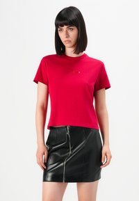 Giovane donna con capelli scuri lisci che indossa una maglietta rossa a maniche corte e una mini gonna di pelle nera con zip frontale.