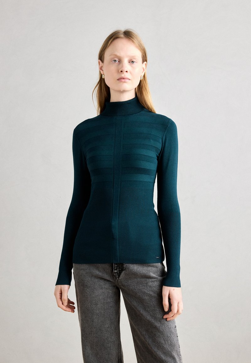 Teal genser med stående hals og ribbet tekstur, med vertikale paneler og lange ermer. Modellen har på seg grå bukser.
