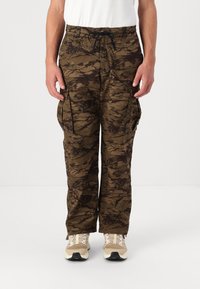 Camo cargo bukser i brune nuancer med store sidelommer, snøre i taljen og afslappet pasform. Båret med lyse sportsko.
