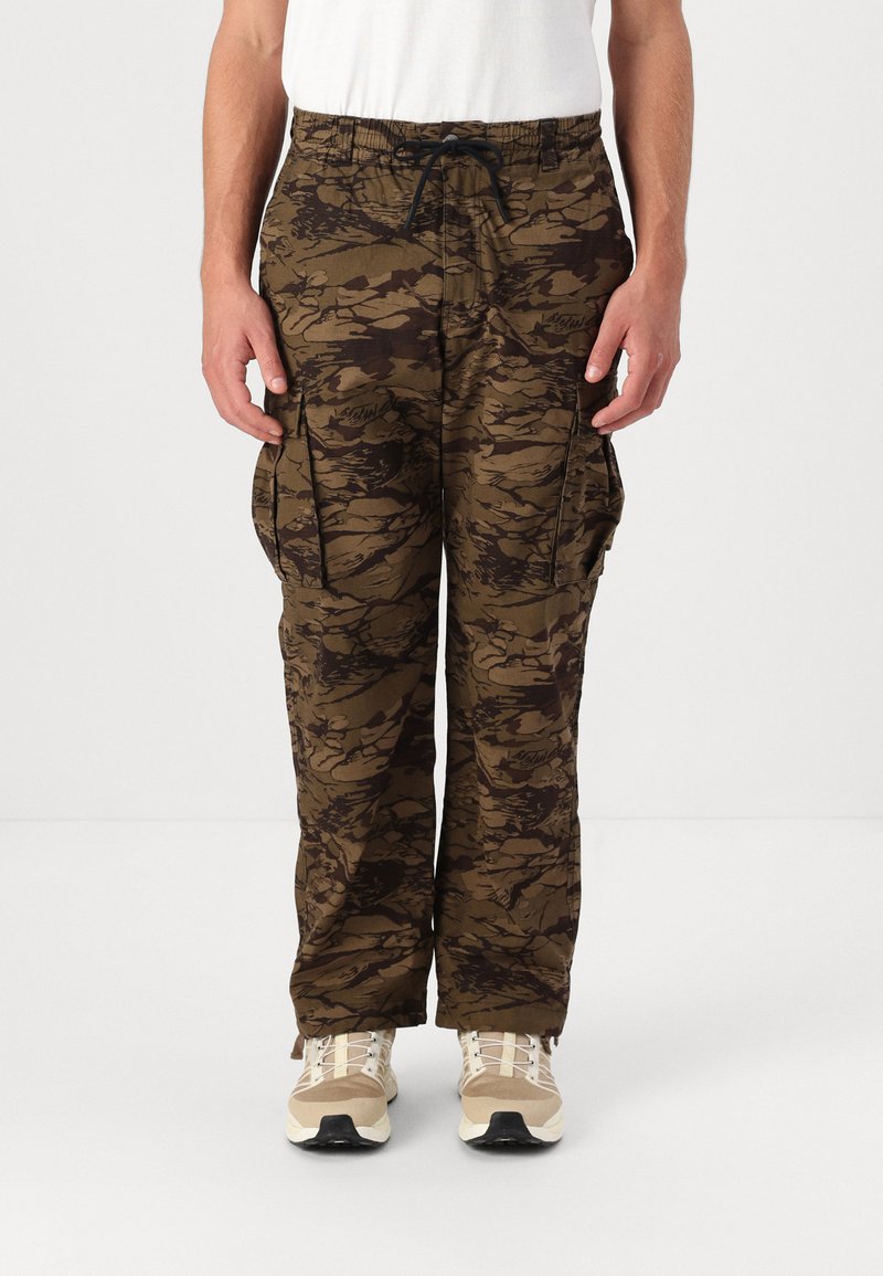 Camo cargo bukser i brune nuancer med store sidelommer, snøre i taljen og afslappet pasform. Båret med lyse sportsko.